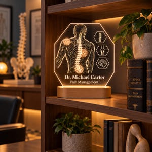 May include: An illuminated hexagonal acrylic sign featuring a line drawing of a human back with highlighted pain points, medical icons, and the text Dr. Michael Carter Pain Management, mounted on a smooth oval wooden base with warm lighting.