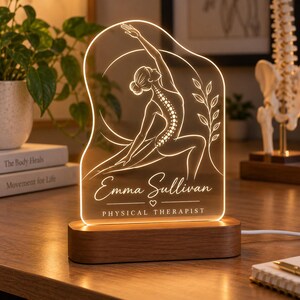 Può includere: Una lampada da scrivania in acrilico illuminata con base in legno, caratterizzata da un disegno a linee incise di una persona in posa yoga con colonna vertebrale visibile, un ramo frondoso e il testo Emma Sullivan PHYSICAL THERAPIST.