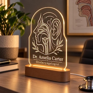 May include: An arched acrylic LED desk lamp with a wooden base, featuring an engraved anatomical illustration of the human ear, nose, and throat. The text reads Dr. Amelia Carter and Otolaryngologist, illuminated by a warm, glowing light.