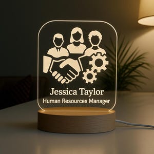 Peut inclure: Lampe de bureau &agrave; LED personnalis&eacute;e avec une base en bois. Elle pr&eacute;sente un graphisme blanc de trois figures, une poign&eacute;e de main et des engrenages. Le nom "Jessica Taylor" et le titre "Human Resources Manager" sont affich&eacute;s.