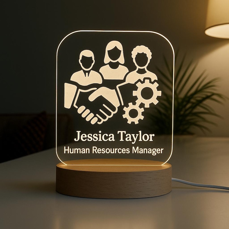 Peut inclure: Lampe de bureau &agrave; LED personnalis&eacute;e avec une base en bois. Elle pr&eacute;sente un graphisme blanc de trois figures, une poign&eacute;e de main et des engrenages. Le nom "Jessica Taylor" et le titre "Human Resources Manager" sont affich&eacute;s.