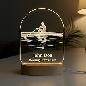 May include: A personalized night light featuring a rowing scene. The design includes a silhouette of a rower on a boat, with the name "John Doe" and the text "Rowing Enthusiast" below. The light sits on a wooden base.