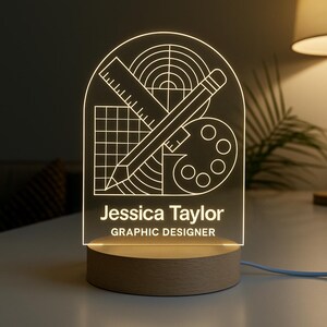 May include: A personalized LED desk lamp with a wooden base. The lamp features a graphic design of a ruler, pencil, palette, and grid, with the name "Jessica Taylor" and the title "Graphic Designer" illuminated in white.