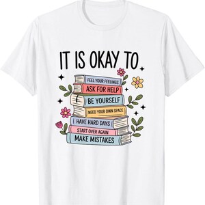 May include: A white short-sleeve t-shirt featuring a graphic of a stack of colorful books with the text IT IS OKAY TO. The book spines are labeled with phrases including FEEL YOUR FEELINGS, ASK FOR HELP, BE YOURSELF, NEED YOUR OWN SPACE, HAVE HARD DAYS, START OVER AGAIN, and MAKE MISTAKES, surrounded by small floral illustrations.