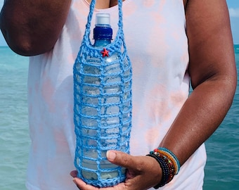 May include: A light blue crocheted water bottle holder with a red star accent. The holder is designed to carry a clear plastic water bottle. The holder has a strap for carrying and is held by a person near the ocean.