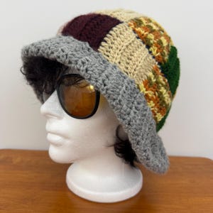 Peut inclure: Un chapeau seau en crochet fait à la main avec un motif patchwork. Le chapeau a un bord gris et une couronne avec des carrés bordeaux, beiges, verts et orange. Le chapeau est présenté sur une tête de mannequin blanche.