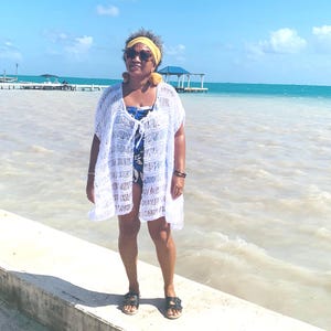 May include: A white, open-weave crochet cover-up with short sleeves. The cover-up is worn over a blue and white patterned swimsuit. The person is wearing a yellow headband, sunglasses, and black sandals. The background features a beach scene with a pier and blue water.
