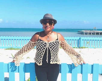 May include: A cream-colored, open-weave crochet top with long sleeves. The top is worn over a black tank top and paired with black pants. The background features a blue fence, ocean, and sky.