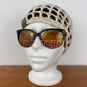 May include: A beige crocheted beanie with a black mesh design and a small brown leather tag. The beanie is paired with black sunglasses with orange-tinted lenses. The word "PATTERN" is printed on the sunglasses.