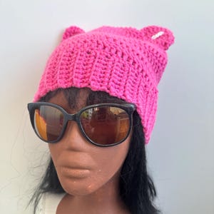 May include: A bright pink, handmade crochet hat with cat ears. The hat is made of thick yarn and has a ribbed texture. The hat is paired with black sunglasses with reflective lenses. The hat is a fun and stylish accessory.