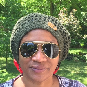 May include: A person wearing a green crocheted beret with a brown leather tag that says "Made with Love". They are also wearing aviator sunglasses.