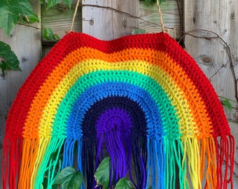 May include: A handmade, rainbow-colored crochet wall hanging. The arched design features bands of red, orange, yellow, green, blue, and purple. The bottom edge has long fringe in matching colors. The wall hanging is suspended by a natural fiber cord.