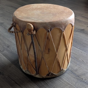 May include: A cylindrical drum with a natural hide drumhead and a light brown wooden body. The drum is approximately 46 cm in diameter and 51 cm tall. It features a woven handle and a wooden beater.