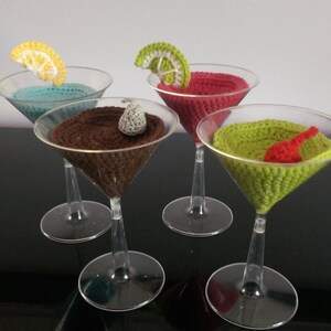 Peut inclure: Quatre verres à martini en crochet avec des boissons et des garnitures de différentes couleurs. Les verres sont en plastique transparent et sont posés sur une surface noire.