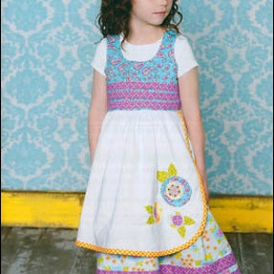 May include: A young girl wearing a white dress with a blue and purple patterned bodice and a matching skirt. She is also wearing a flower headband and a pair of black shoes. The dress has a yellow trim and a flower applique. The text "kati cupcake patterns" and "Designs by nayhamberlin" is visible in the top left corner of the image. The text "Ruth & Ruby" is visible in the bottom right corner of the image.