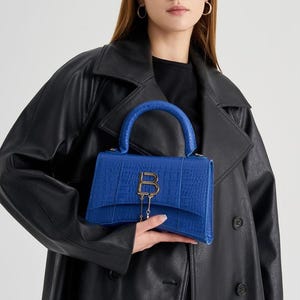 May include: A vibrant blue handbag with a textured crocodile-embossed finish, featuring a structured silhouette, a single top handle, and a prominent gold-toned letter B logo on the front flap with a delicate dangling gold chain detail.