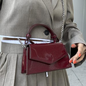 Peut inclure: Un sac &agrave; main en cuir verni bordeaux avec une bandouli&egrave;re en cha&icirc;ne argent&eacute;e et une poign&eacute;e sup&eacute;rieure. Le sac a une forme rectangulaire avec une fermeture &agrave; rabat. Le sac est port&eacute; devant un blazer et une jupe gris.
