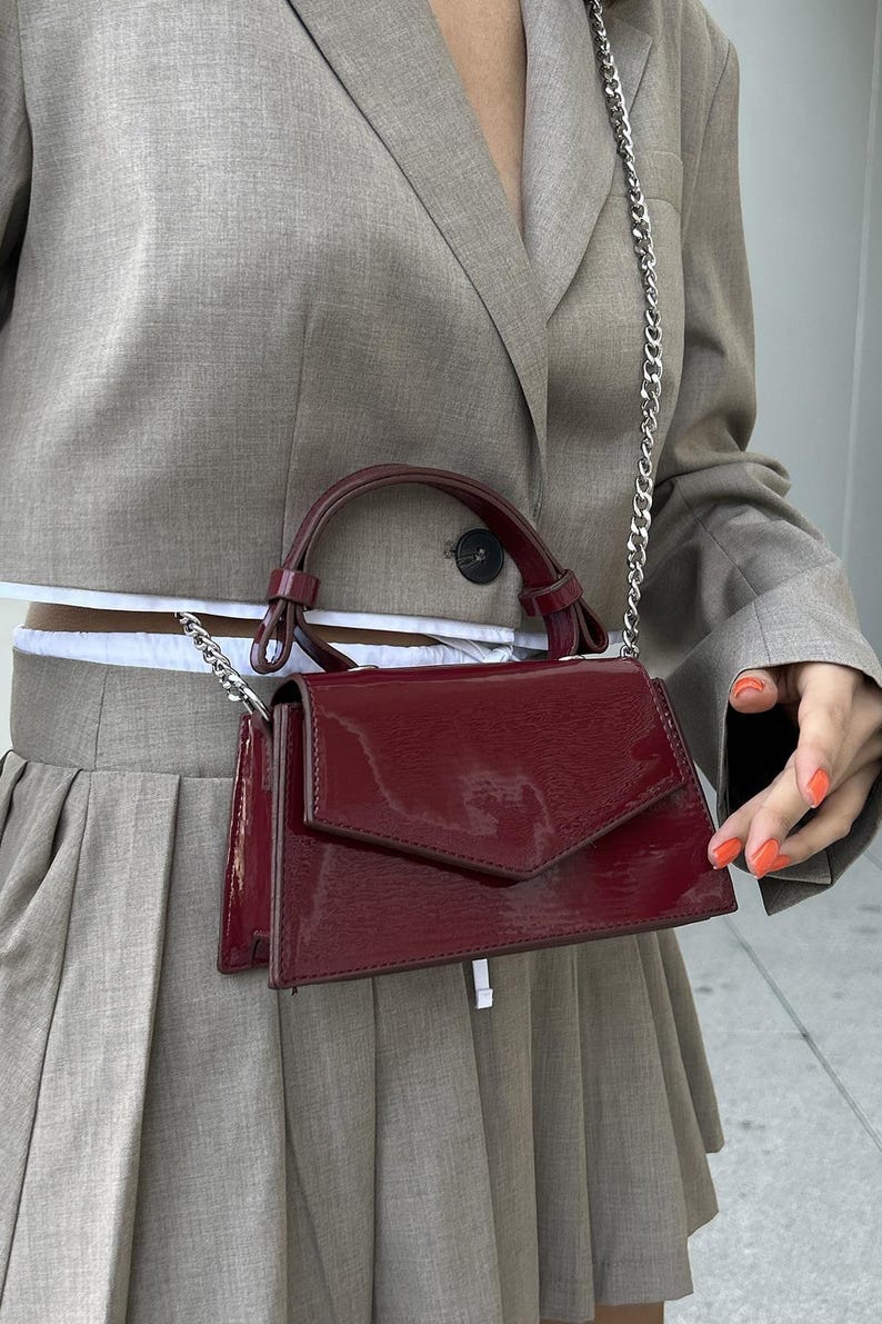 Peut inclure: Un sac &agrave; main en cuir verni bordeaux avec une bandouli&egrave;re en cha&icirc;ne argent&eacute;e et une poign&eacute;e sup&eacute;rieure. Le sac a une forme rectangulaire avec une fermeture &agrave; rabat. Le sac est port&eacute; devant un blazer et une jupe gris.