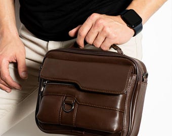 Puede incluir: Un maletín de cuero marrón con asa superior, solapa delantera y bolsillo con cremallera. El bolso tiene forma rectangular y un detalle de anilla en D metálica. El maletín lo sostiene una persona que lleva una camisa negra y un reloj.