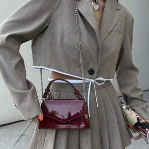 Peut inclure: Un ensemble &eacute;l&eacute;gant compos&eacute; d'un blazer court beige, d'une jupe pliss&eacute;e assortie et d'un sac &agrave; main bordeaux brillant avec une cha&icirc;ne argent&eacute;e. L'ensemble est compl&eacute;t&eacute; par un choker noir et un pendentif c&oelig;ur argent&eacute;.