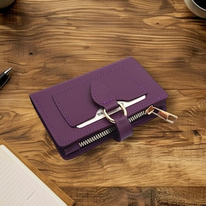 Może przedstawiać: Fioletowy portfel ze złotym zamkiem błyskawicznym i miejscem na karty. Portfel wykonany jest z gładkiego materiału i posiada pasek z zapięciem na złoty pierścień. Portfel leży na drewnianej powierzchni.