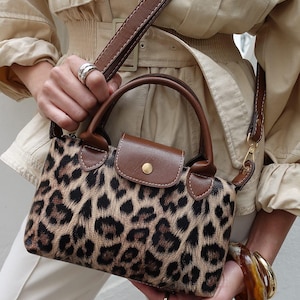 May include: A small handbag with a leopard print pattern. The bag has brown leather accents on the handles, flap, and strap. A gold-colored clasp and button secure the flap. The bag is held by a person wearing a beige jacket and white pants.