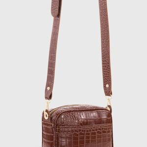 May include: A brown rectangular crossbody bag with a crocodile-embossed texture. The bag features a gold-tone zipper closure, matching gold-tone hardware, and an adjustable shoulder strap with a buckle, all finished in a consistent brown crocodile pattern.