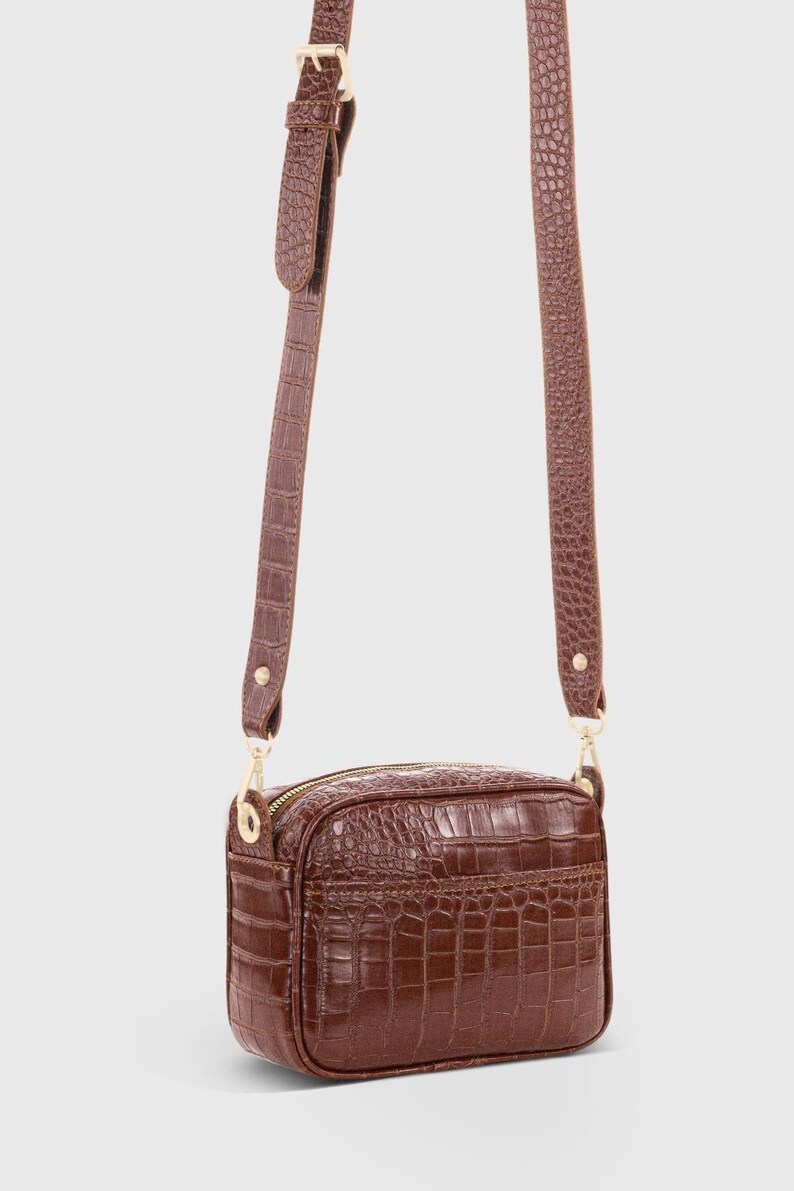 May include: A brown rectangular crossbody bag with a crocodile-embossed texture. The bag features a gold-tone zipper closure, matching gold-tone hardware, and an adjustable shoulder strap with a buckle, all finished in a consistent brown crocodile pattern.