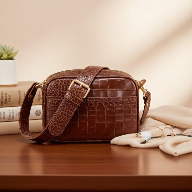 May include: A brown rectangular crossbody bag with a crocodile-embossed texture, featuring a matching adjustable strap with a gold-tone metal buckle, a top zipper closure, and gold-tone hardware accents on the sides of the bag.