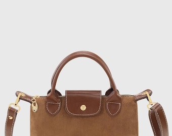 May include: A brown suede handbag with a top handle and a detachable shoulder strap. The bag features a flap closure with a gold-coloured button and gold-coloured hardware. The bag's dimensions are approximately 25 cm wide, 20 cm high, and 10 cm deep.