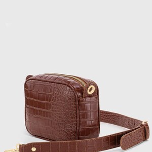 May include: A brown crossbody bag featuring a textured crocodile-embossed pattern. The rectangular bag includes a gold-tone zipper closure, matching gold-tone hardware, and an adjustable shoulder strap with a gold-tone metal clasp and stud detail.