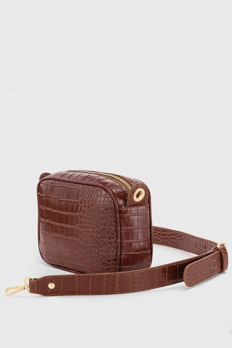 May include: A brown crossbody bag featuring a textured crocodile-embossed pattern. The rectangular bag includes a gold-tone zipper closure, matching gold-tone hardware, and an adjustable shoulder strap with a gold-tone metal clasp and stud detail.