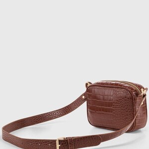 May include: A brown belt bag featuring a textured crocodile-embossed pattern. The rectangular bag has a top zipper closure and is attached to an adjustable matching strap with a gold-toned metal buckle and hardware, creating a cohesive look.