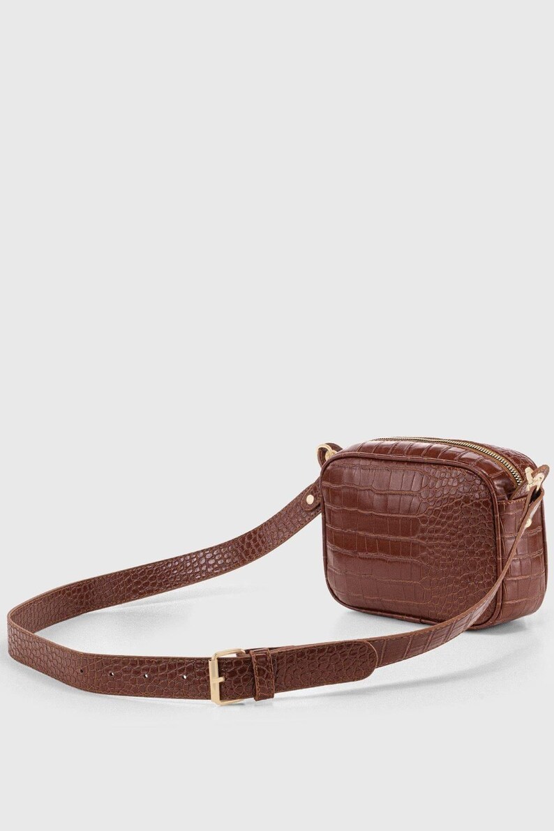 May include: A brown belt bag featuring a textured crocodile-embossed pattern. The rectangular bag has a top zipper closure and is attached to an adjustable matching strap with a gold-toned metal buckle and hardware, creating a cohesive look.