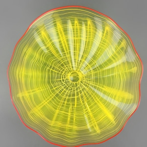 May include: A yellow glass wall sculpture with a red rim. The glass has a wavy, rippled texture and a circular pattern.