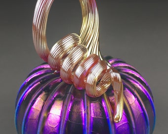 May include: A glass pumpkin with a iridescent purple and blue finish. The stem is a swirl of gold and pink glass.
