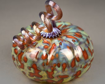 Puede incluir: Una calabaza de vidrio con un diseño de espiral en tonos verdes, azules y naranjas. La calabaza tiene un tallo iridiscente marrón y morado.
