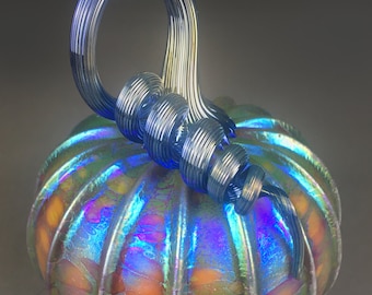 May include: A glass pumpkin with a blue iridescent finish and a swirling blue glass stem. The pumpkin has a ribbed texture and is decorated with small, iridescent spots.