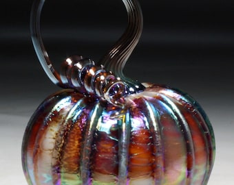 May include: A handcrafted glass pumpkin with a swirling stem and handle. The pumpkin has a ribbed design and iridescent finish, displaying hues of orange, purple, and green. The stem and handle are clear glass.