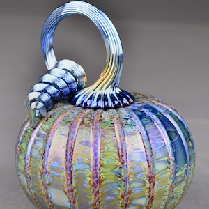 May include: A glass pumpkin with a blue and gold iridescent finish. The pumpkin has a ribbed texture and a twisted stem.