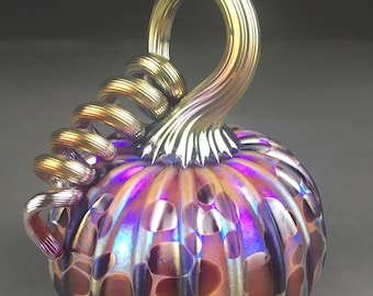 May include: A decorative glass pumpkin with a swirling stem. The pumpkin has a textured surface with a pattern of brown and purple spots. The stem is gold and the body of the pumpkin has iridescent colors.
