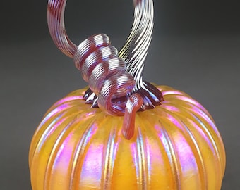 May include: A glass pumpkin with a ribbed surface and iridescent finish. The pumpkin is a vibrant yellow with pink and purple highlights. The stem is a twisted, iridescent purple glass.