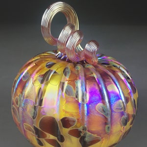 May include: A glass pumpkin with a swirl design in iridescent yellow, orange, and purple hues. The pumpkin has a brown and yellow spotted pattern and a curled, pink glass stem.
