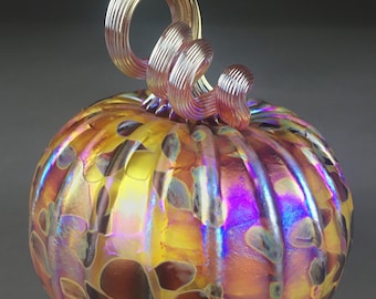 May include: A glass pumpkin with a swirl design in iridescent yellow, orange, and purple hues. The pumpkin has a brown and yellow spotted pattern and a curled, pink glass stem.