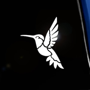 May include: White hummingbird decal in flight, affixed to a dark surface. The decal features detailed wing and tail feather designs. The Seward Street Studios logo is in the upper left corner and the copyright is in the lower right corner.