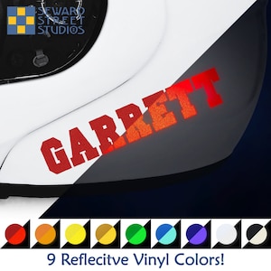 May include: A red reflective vinyl decal with the name "GARRETT" in a bold font. The decal is shown on a white motorcycle helmet. The image also shows nine different reflective vinyl colors.