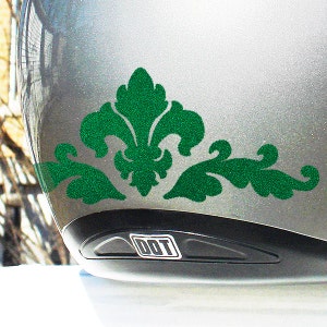 May include: A silver motorcycle helmet with a green glitter decal of a stylized floral design. The decal is on the back of the helmet, near the bottom. The helmet has a black DOT sticker on the back.