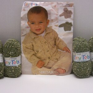 May include: Four balls of green and white yarn with a label that reads "Sirdar Snuggly Tiny Tots" and a knitting pattern for a baby cardigan. The pattern is titled "Tiny Tots DK" and has a picture of a baby wearing the cardigan.