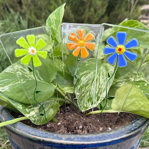 May include: Three glass flower garden stakes with colorful daisy designs. The flowers are green, orange, and blue, each with a different colored center. The stakes are placed in a potted plant with green and white variegated leaves.