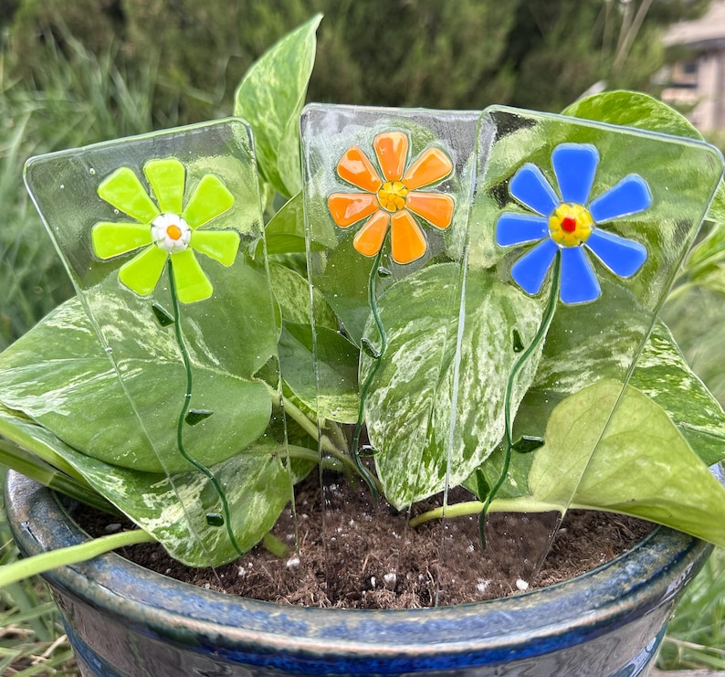 May include: Three glass flower garden stakes with colorful daisy designs. The flowers are green, orange, and blue, each with a different colored center. The stakes are placed in a potted plant with green and white variegated leaves.